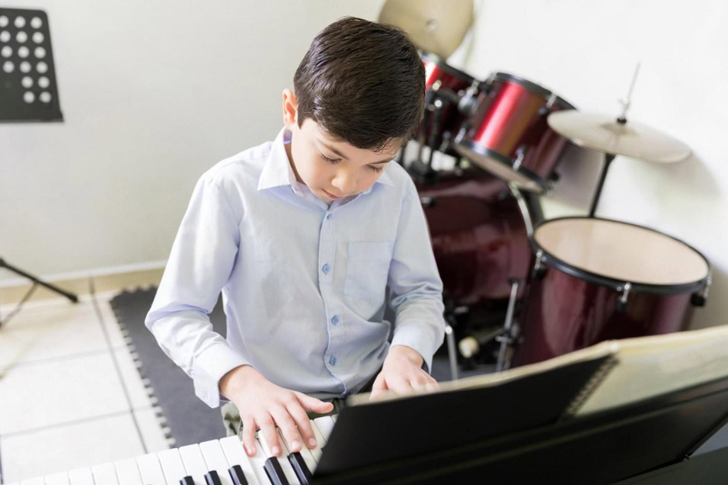 Young boy in a blue shirt playing an electric piano with a drum kit in the background during a music lesson